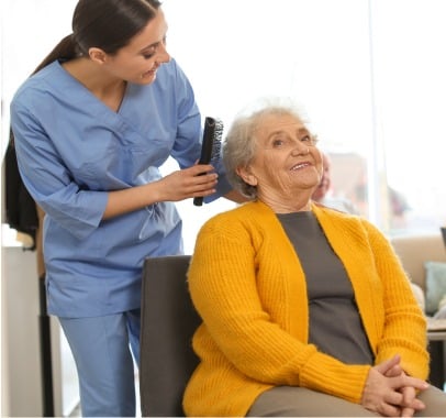 A young nurse in blue scrubs is brushing the hair of an elderly woman wearing a yellow cardigan. The woman is sitting in a chair and smiling, while the nurse happily chats with her. The setting appears to be a bright, comfortable room at an adult day care center in Burlington County NJ.