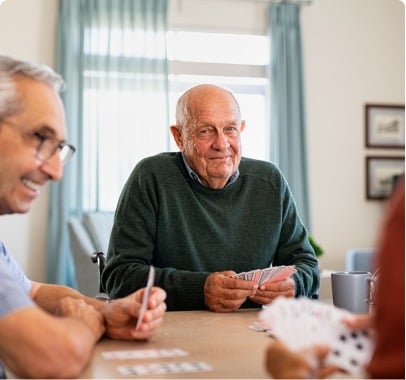 An elderly man with a green sweater and glasses sits at a table, holding playing cards and smiling. He is surrounded by other people also playing cards. The room has large windows with sheer curtains, letting in natural light, providing a welcoming atmosphere for those enjoying senior care in Burlington County NJ.