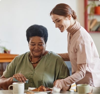 A caregiver in a light pink uniform is smiling and assisting an elderly woman in a green top at an adult day care facility in Burlington County NJ. The elderly woman sits at a table with a plate of food and a cup, while the caregiver stands beside her, placing a hand gently on her shoulder.