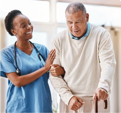 A nurse in blue scrubs and a stethoscope around her neck is assisting an elderly man, who is using a cane, at an adult day care facility in Burlington County NJ. The man, wearing a light sweater and a serious expression, is being supported by the smiling nurse holding his arm.