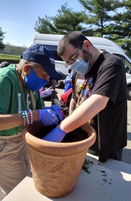 Two people at an adult day care facility in Burlington County NJ are planting in a large terracotta pot outdoors. One, wearing a blue cap and green shirt, assists the other who is donning glasses, a mask, and a black shirt. They both have on protective gloves while a vehicle is visible in the background.