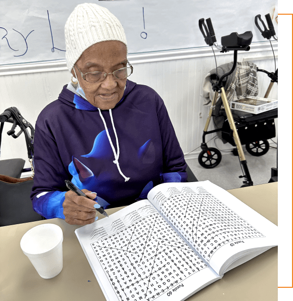 An elderly person wearing a white knit hat, glasses, and a blue hoodie is seated at a table working on a word search puzzle with a pen. There's a white cup on the table and a walker in the background, reminiscent of scenes from an adult day care center in Burlington County NJ.