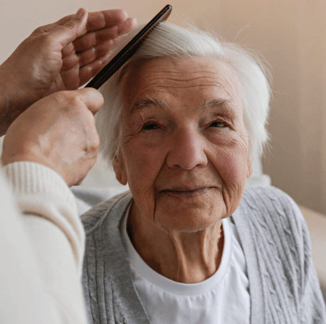 An elderly person with short white hair is having their hair gently combed by another person. The elderly person is smiling slightly and looking at the camera. The setting appears to be an adult day care center in Burlington County, NJ, with soft lighting and a calm atmosphere.