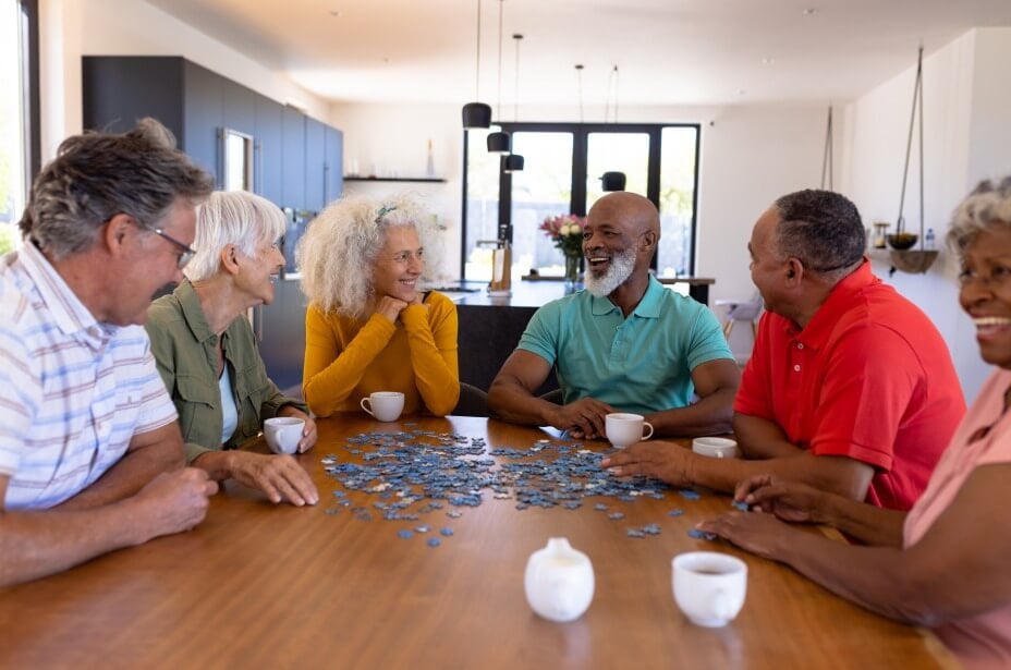 A group of six older adults, three men and three women, sit around a wooden table, smiling and conversing while working on a jigsaw puzzle in an adult day care facility in Burlington County NJ. Cups of coffee are on the table, and natural light fills the bright, modern kitchen through large windows.
