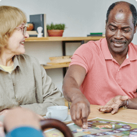 Two individuals sit at a table, engaged in a game or activity at an adult day care facility in Burlington County NJ. The person on the left, wearing glasses, a beige shirt, and smiling, looks at the other person who is wearing a pink polo shirt and is actively pointing at the game or activity on the table.