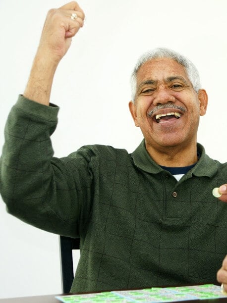 A smiling older man with gray hair and a mustache raises his arm in celebration. He is seated at a table with a bingo card in front of him. He wears a dark green, checkered sweater.
