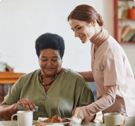 A caregiver in a pink uniform assists an elderly woman with short black hair, who is seated at a table in an adult day care facility in Burlington County, NJ. The woman, wearing a green blouse, reaches for some food in front of her as they enjoy the warmth of the room.