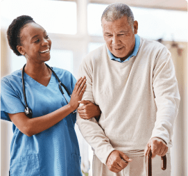 A healthcare worker in blue scrubs, wearing a stethoscope, is smiling and assisting an elderly man who is using a cane. The scene appears cheerful as they walk together in a bright, well-lit environment at an adult day care center in Burlington County NJ.