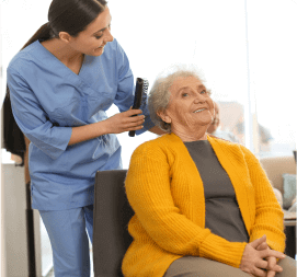 A caregiver in blue scrubs brushes the hair of a smiling elderly woman in a yellow cardigan at an adult day care center in Burlington County NJ. They are indoors, and the elderly woman is seated while the caregiver stands behind her, interacting warmly.