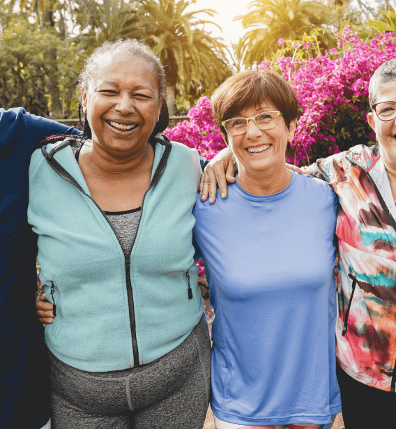 Four women are standing outdoors with their arms around each other, smiling. They are dressed casually in activewear, and behind them is a vibrant backdrop of pink flowers and lush greenery. It appears to be a sunny day at an adult day care center in Burlington County NJ.