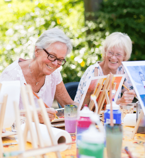 Two elderly women seated outdoors at an adult day care in Burlington County NJ are joyfully painting together. They are surrounded by easels, paintbrushes, and colorful paint bottles. Both are smiling and appear to be enjoying the creative activity on a sunny day with trees and greenery in the background.