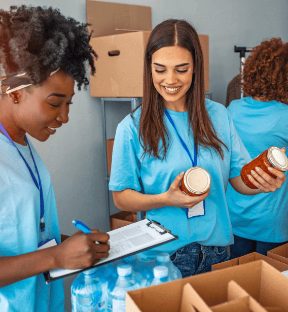 Two women wearing blue shirts and lanyards are sorting and organizing jars of food, with one writing on a clipboard. They are surrounded by cardboard boxes and water bottles, participating in a community service activity at an adult day care facility in Burlington County NJ.