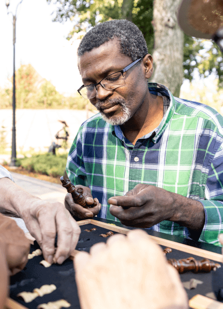 A man with glasses and graying hair wearing a green checked shirt is playing chess outdoors at a senior care facility near Camden County, NJ. His attention is focused on the chess pieces as he makes his move. Two other people's hands are visible in the foreground, also interacting with the chess pieces.