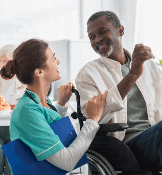 A nurse in a teal uniform smiles and talks with a man sitting in a wheelchair. The man, wearing a white shirt, gestures with his hand while looking at the nurse. Both appear happy and engaged in their conversation, showcasing the warmth of senior care in Burlington County NJ.